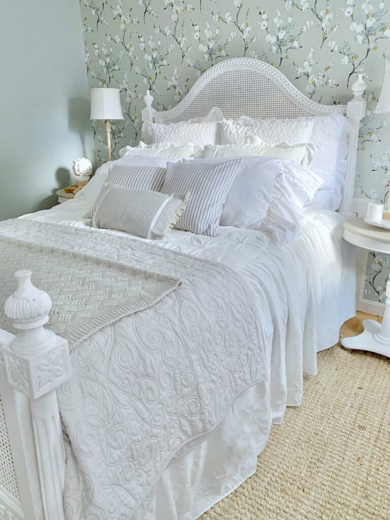 Sideview of a white bed with caning. lot of flowy bedspreads and coverlets with pillows.there is a floral print wall paper accent wall behind the bed. 