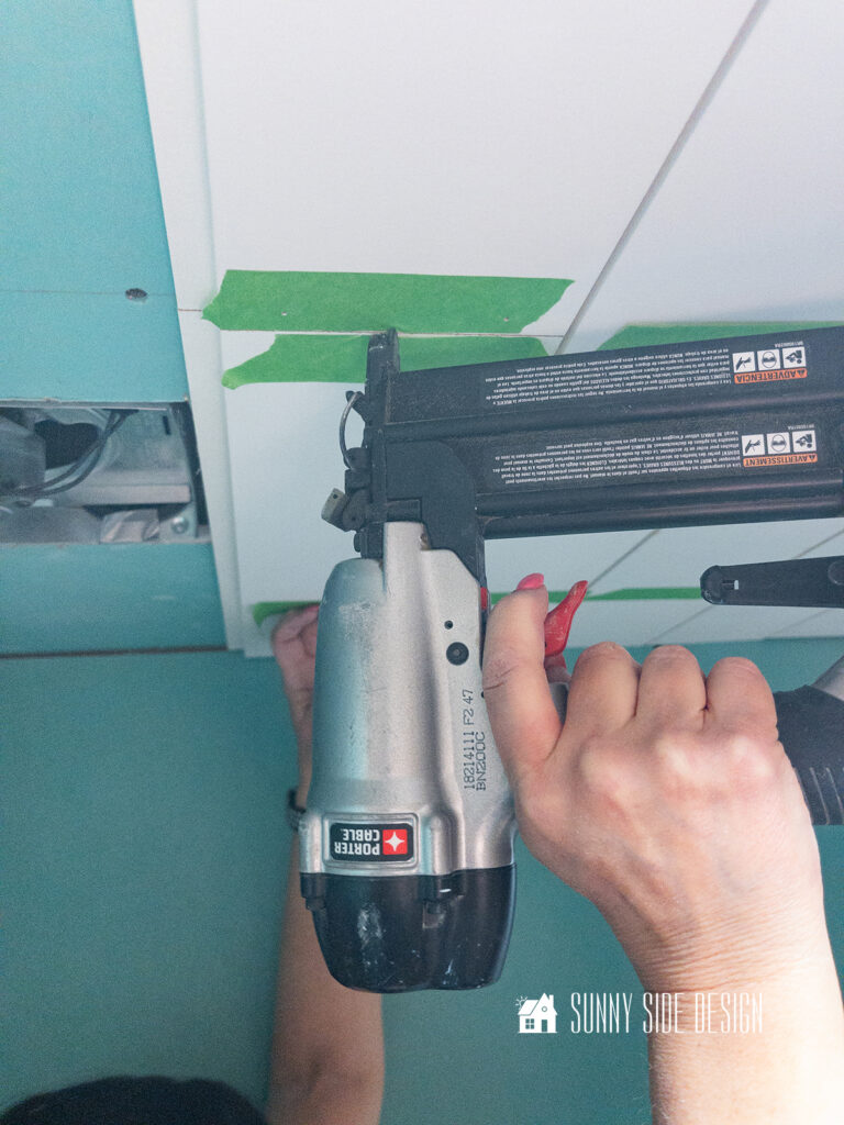 Woman shoots brad nails through painters tape into shiplap on bathroom ceiling.