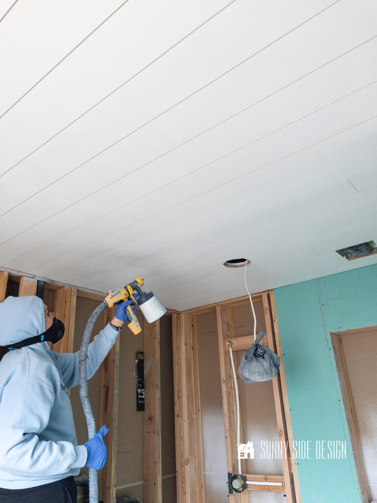 Woman using a paint sprayer painting the shiplap ceiling in the bathroom.