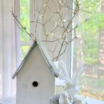 A large wooden white bird house in front of a white pitcher of faux cherry blossom branches.